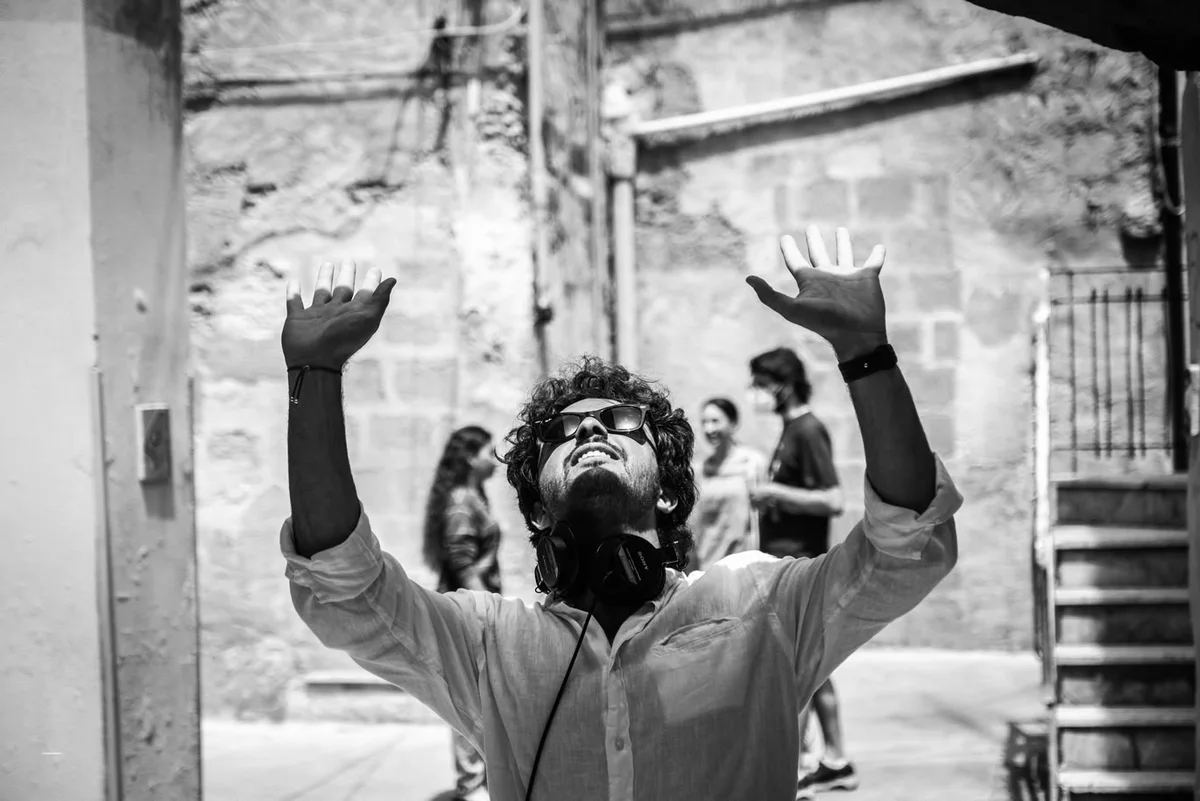 Fabio Schifilliti on set in a sunlit alley, headphones around his neck, both arms raised.
